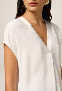 Blusa blanca ligera con diseño de cuello en V y mangas cortas enrolladas; textura suave y estilo minimalista; sin patrones visibles ni hardware.