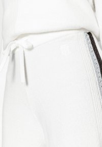 White fabric joggers with a soft texture, featuring a drawstring waistband and contrasting side stripes in darker tones. Embossed logo detail.