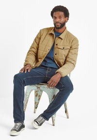 Tan quilted jacket with a front pocket, worn over a blue shirt. Dark blue jeans and black sneakers with white soles, seated on a rusty metal chair.