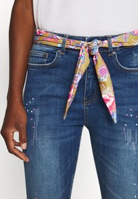 Tmavě modré džíny s květinovým pásem z látky v odstínech růžové a žluté, s ozdobami na levém stehně.