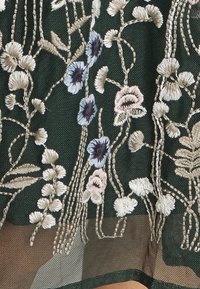 Dunkelgrüner Stoff mit aufwendiger floraler Stickerei in Beige, Pink und Blau. Das Design umfasst verschiedene Blumen und strukturierte Details.