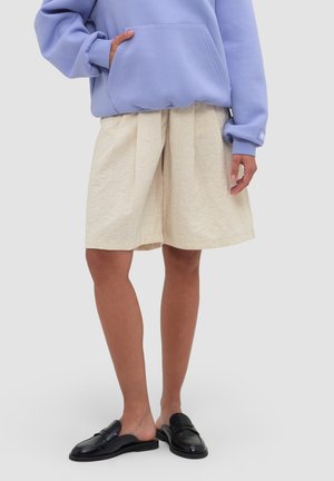 Persoon draagt beige geplooide shorts, een lavendelkleurige hoodie met hand in de zak en zwarte slip-on loafers tegen een eentonige achtergrond.