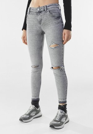 Kvinde iført lysegrå revnede skinny jeans, sorte sokker, grå og hvide sneakers samt sort langærmet cropped top, stående på hvidt gulv.