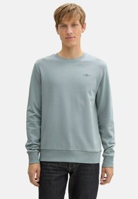 Hellblauer Sweatshirt aus glattem Stoff mit langen Ärmeln, geripptem Kragen und Saum, ausgestattet mit einem kleinen Logo und Datumsangabe auf der Brust.