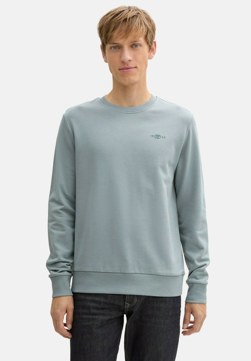 Hellblauer Sweatshirt aus glattem Stoff mit langen Ärmeln, geripptem Kragen und Saum, ausgestattet mit einem kleinen Logo und Datumsangabe auf der Brust.