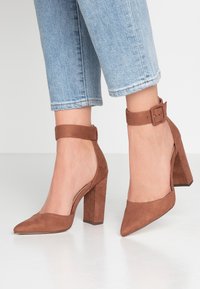 Brown suede high-heeled shoes with pointed toes and ankle straps. Features a chunky heel and smooth texture. Worn with cropped denim.