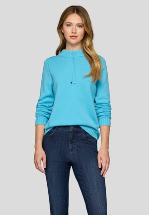 Frau trägt einen hellblauen Langarmpullover mit hohem Kragen und dunkelblauen Jeans, steht vor einem schlichten grauen Hintergrund.