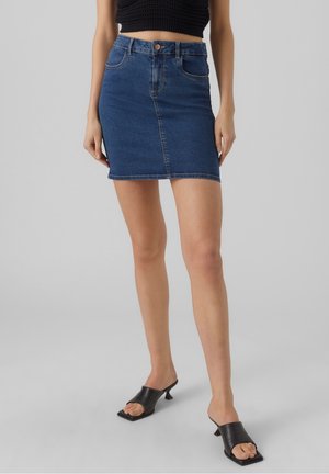 Kvinde iført en mørkeblå denim mininederdel, sort kortstrikket top og sorte åbne kitten heel mules, der står mod en ensfarvet baggrund.