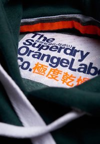 Sudadera verde con cordones blancos, que presenta una etiqueta texturizada con la inscripción "The Superdry OrangeLabel" en texto azul y naranja.