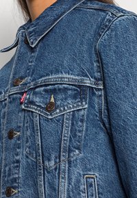 Sininen farkkutakki, jossa on kaulus, etutaskut ja ruskeat metallinapit. Haalistunut pinta ja keltaiset ompeluyksityiskohdat.