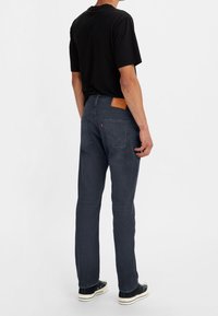Jeans de mezclilla azul oscuro con un corte recto, presentando un parche de cuero en la cintura y dos bolsillos traseros, combinados con una camiseta negra y zapatillas.