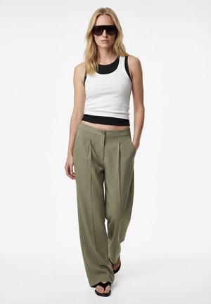 Blondynka w czarnych okularach przeciwsłonecznych, warstwowych czarno-białych bezrękawnikach, oliwkowozielonych spodniach z szerokimi nogawkami i czarnych klapkach, idąca do przodu.