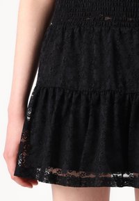 Svart blondekjole med smokkebodice og lagdelt volangnederdel. Stoffet har et blomsterdesign med gjennomsiktige detaljer.