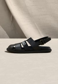 Sandale pour homme en cuir noir avec pointe fermée, plusieurs lanières dont une avec un texte blanc, et une boucle ajustable au talon.