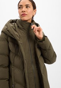 Abrigo acolchado verde oliva con cuello alto, cierre de cremallera y tejido texturizado. Costuras visibles y capucha realzan el diseño.