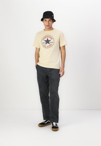 Camiseta de color beige claro con un gráfico circular en rojo y azul, sombrero tipo cubo negro, pantalones de pana gris y zapatillas negras con detalles blancos.
