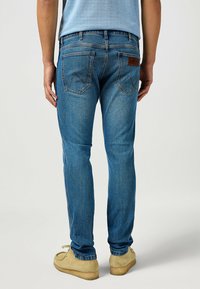 Blå denimjeans med rak skärning, med två bakfickor och en brun läderlapp; kombinerade med beige mockasiner.