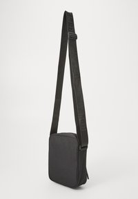 Black fabric crossbody bag with a square shape, textured surface, and an adjustable strap featuring embossed branding. Zipper closure on the side.