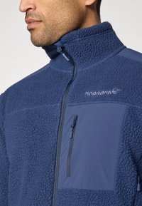 Veste en polaire marine avec col haut, poche poitrine zippée et accents de tissu lisse contrastants. Présente un tissu texturé sur l'ensemble.