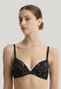 Soutien-gorge en dentelle noire avec un design triangulaire, présentant des motifs floraux et des détails délicats, maintenu par des fines bretelles ajustables.