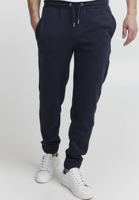 Navy sweatpants with an elastic waistband and drawstring, featuring side pockets and tapered cuffs. Paired with white sneakers.