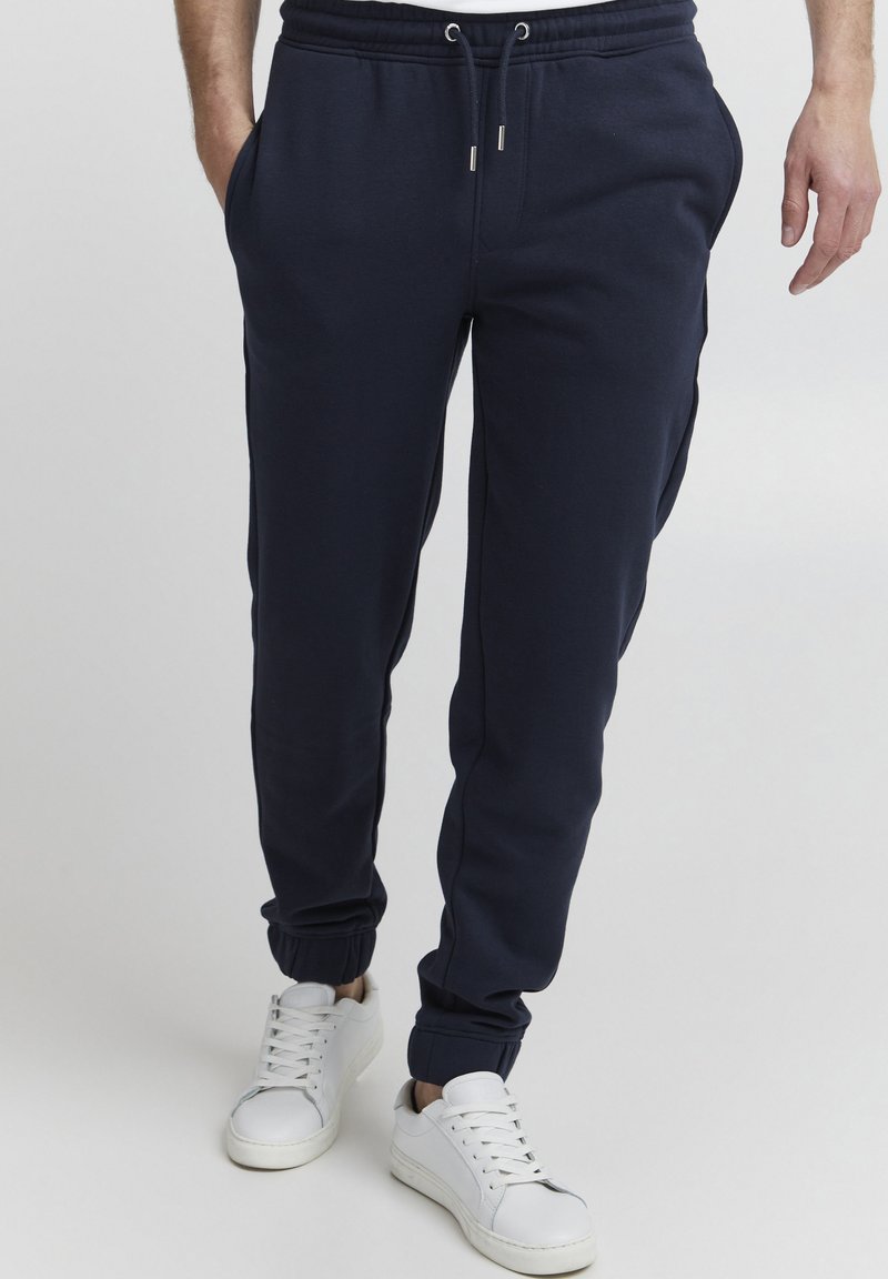 Navy sweatpants with an elastic waistband and drawstring, featuring side pockets and tapered cuffs. Paired with white sneakers.