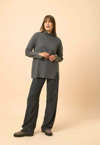 Grau gerippter Strickpullover mit Rollkragen und seitlichen Schlitzen, kombiniert mit lockeren, dunklen Hosen und grauen Schuhen, vor einem hellen Hintergrund.