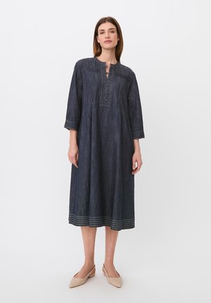 Kvinde står iført en mørkeblå, langærmet, midi-lang kjole med knaplukning foran og beige lavhælede sko på en hvid baggrund.