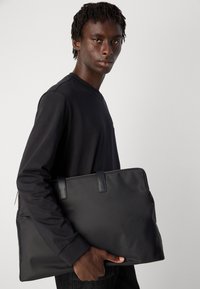 Ung mann i svart langermet skjorte som holder en stor svart glidelåse-tote bag mot en enkel lys bakgrunn.