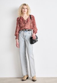 Rood gemusterde blouse met ruffles, hoogsluitende lichtblauwe spijkerbroek en beige enkelboots. Zwarte handtas met een kettingband.