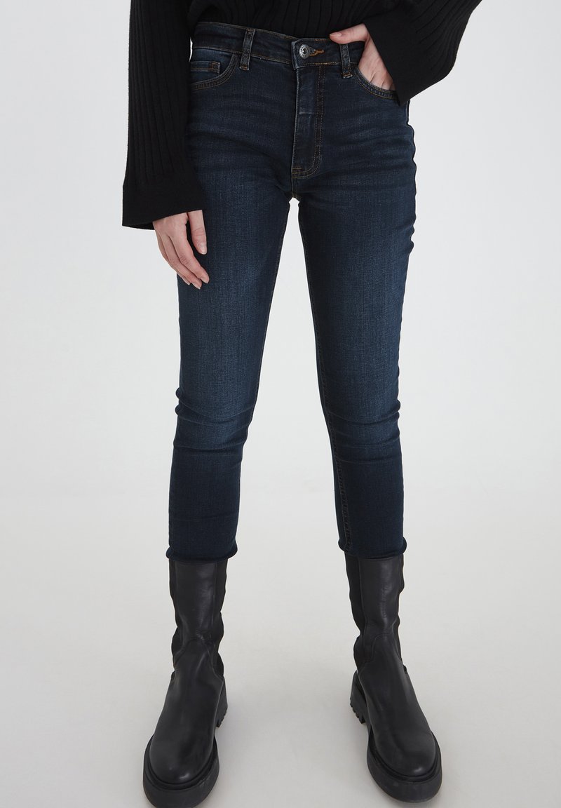 Jeans skinny de color azul oscuro con cintura alta, hechos de tela de mezclilla. Combinados con botas negras de tobillo. Con cinco bolsillos y cierre de botón.