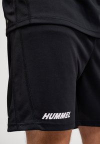 Gros plan d'une personne portant un short de sport noir avec un logo blanc "HUMMEL" sur l'ourlet inférieur.