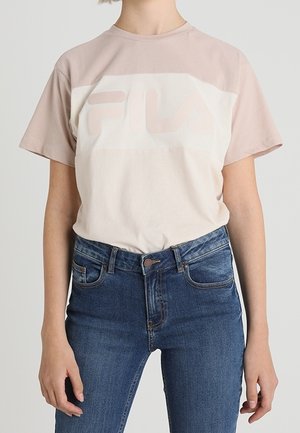 Person wearing a beige and light pink FILA t-shirt tucked into high-waisted dark blue jeans against a plain background.