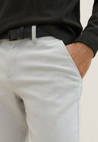 Lichtgrijze textuurbroek met een getailleerde snit, gedragen met een donkere trui en een zwarte riem. Zak zichtbaar aan de zijkant.