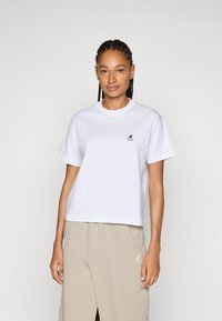 Valge puuvillane T-särk lühikeste varrukatega, ümmargune kaelus ja väike must logo vasakul rinnal. Veidi vaba, juhuslik sobivus.