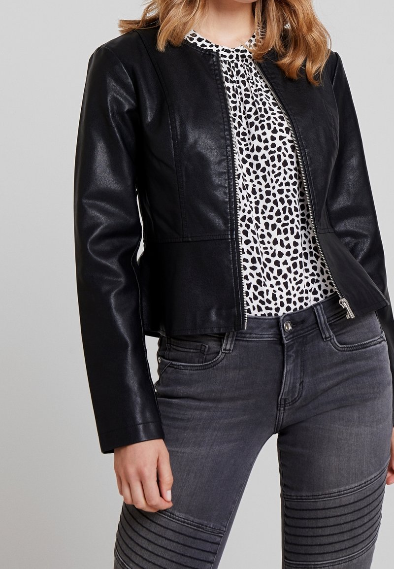 Femme portant une veste en cuir noire sur un chemisier blanc et noir à pois et un jean gris foncé avec des détails côtelés sur les cuisses.