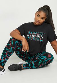Musta t-paita, jossa on rohkea teksti "#OLE ITSESI" yhdistetty värikkäisiin, kuvioituihin legginsseihin mustassa, tealissa ja burgundissa. Mustat urheilukengät.