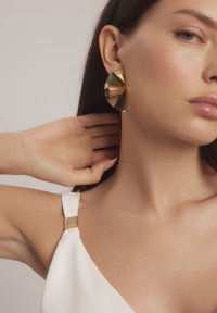 Złote kolczyki o geometrycznym wzorze, w kształcie przypominającym wachlarz, z wypolerowaną powierzchnią. Modelka nosi biały top z złotym akcentem.
