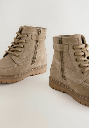 Stivali alla caviglia in suede beige con superficie texturizzata, punta rotonda e chiusura con lacci. Zip laterale per una facile calzata e suole in gomma con grip.