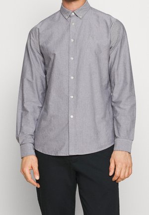 Man wearing a buttoned-up, long-sleeve, light gray shirt with a collar and small embroidered logo on the chest, paired with black pants.