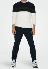 Pletený svetr v námořnické modři, krémové a tmavě zelené pruhy; spárován s černými kalhotami a barevnými sportovními botami.