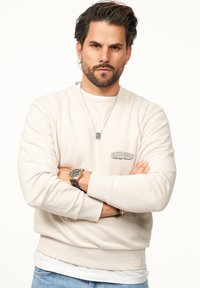 Helles beiges Sweatshirt mit rundem Hals, langen Ärmeln und Logodetail. Über einem weißen Hemd getragen, mit einer silbernen Halskette und Uhr ergänzt.