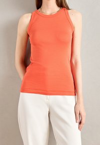 Top in cotone arancione senza maniche con un design aderente, scollo rotondo e una texture liscia, abbinato a pantaloni bianchi.