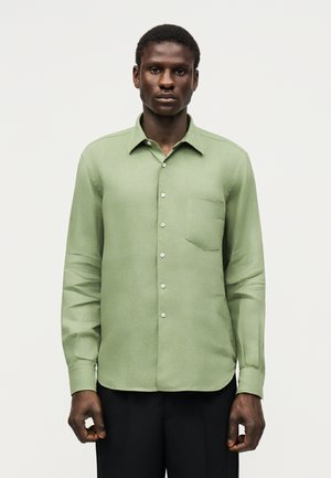 Άνδρας που φορά ένα ανοιχτοπράσινο πουκάμισο με μακρύ μανίκι και τσέπη στο στήθος, που στέκεται μπροστά σε απλό ανοιχτό φόντο.