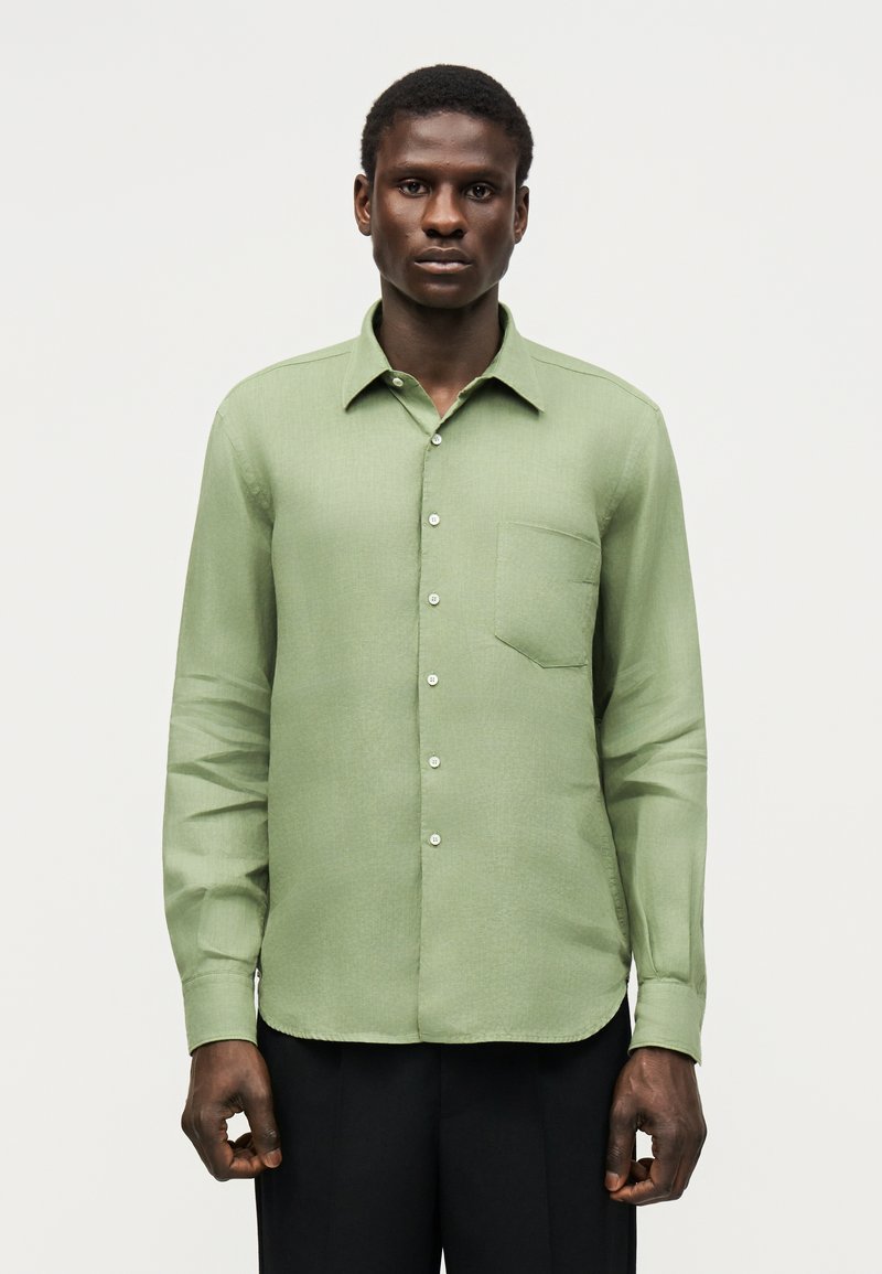 Άνδρας που φορά ένα ανοιχτοπράσινο πουκάμισο με μακρύ μανίκι και τσέπη στο στήθος, που στέκεται μπροστά σε απλό ανοιχτό φόντο.