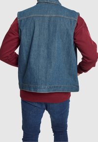 Jeansväst i ljusblått, med en klassisk design och utan ärmar. Bärs över en vinröd sweatshirts och matchas med blå jeans.