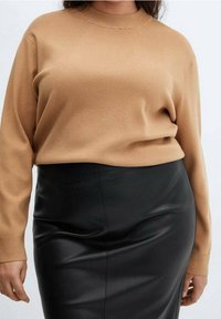 Femme portant un pull à manches longues beige rentré dans une jupe en cuir noire taille haute, debout devant un fond clair uni.
