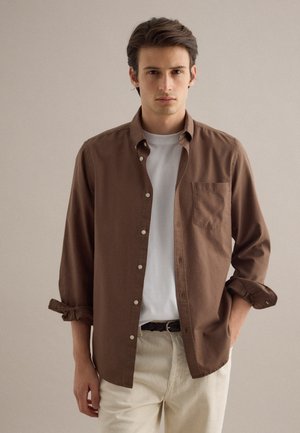 Junger Mann mit dunklen Haaren trägt ein braunes, geknöpftes Hemd über einem weißen T-Shirt und beigefarbene Hosen, steht vor einem schlichten hellen Hintergrund.