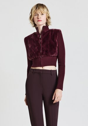 Maglione corto bordeaux con fronte in pelliccia ecologica morbida e maniche a coste in maglia, abbinato a pantaloni scuri su misura con chiusura a zip.