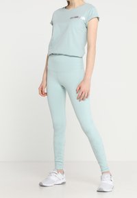 Žena v světle modrých legínách s vysokým páskem a jemným písmenkovým vzorem na dolních nohavicích, světle modrém tričku s ohrnutými rukávy a bílých teniskách.